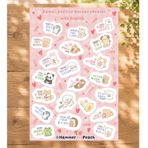 May include: A sticker sheet with a light pink plaid background featuring various cartoon animals including a penguin, cat, dog, bear, and frog. Each sticker includes Korean text and English translations. The top reads kawaii positive korean phrases with English and the bottom reads Hammer and Peach.