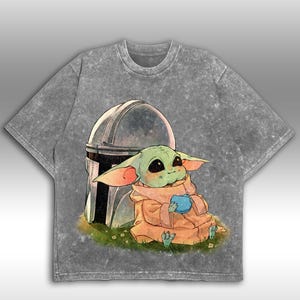 May include: A grey acid wash t-shirt with a graphic of a green alien character holding a blue object, with a silver helmet behind it. The character sits on grass with flowers. The t-shirt has a crew neck and short sleeves.