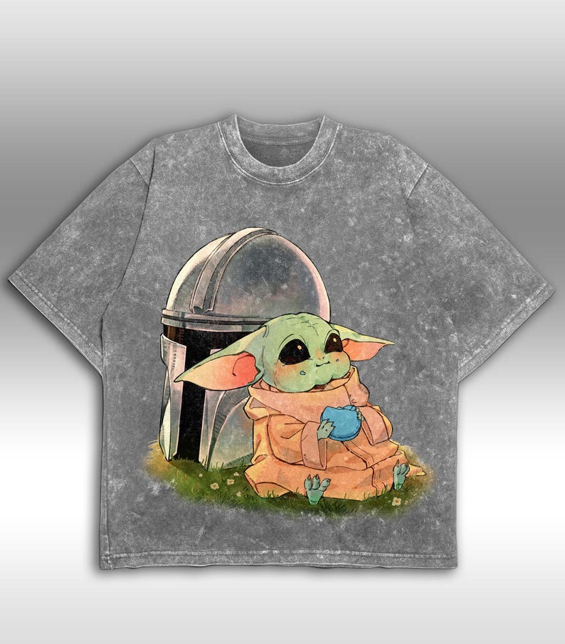 May include: A grey acid wash t-shirt with a graphic of a green alien character holding a blue object, with a silver helmet behind it. The character sits on grass with flowers. The t-shirt has a crew neck and short sleeves.