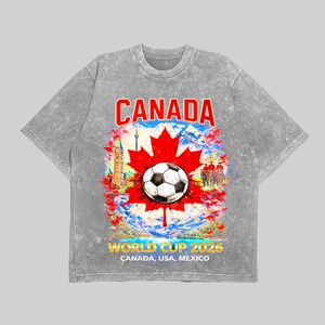 May include: A gray t-shirt featuring a graphic design for the 2026 World Cup. The design includes a soccer ball, a red maple leaf, and the word "CANADA" in red. The text "WORLD CUP 2026 CANADA, USA, MEXICO" is also included.