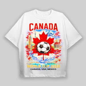 May include: White t-shirt featuring a soccer ball on a red maple leaf, with the word "CANADA" in red above. The shirt also includes the text "WORLD CUP 2026 CANADA, USA, MEXICO". The design incorporates Canadian landmarks and scenery.