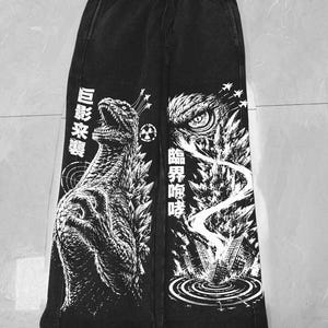 May include: Black sweatpants featuring a white graphic print of a monster on the left leg and a close-up of a monster eye over a city on the right leg. The design includes Japanese text, a radiation symbol, and small airplanes in the sky.
