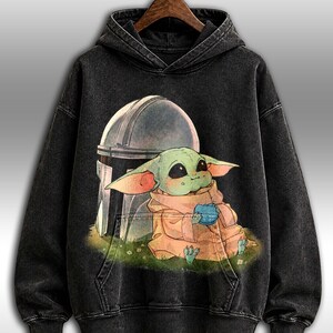 May include: Dark grey hooded sweatshirt with a graphic of Grogu, or Baby Yoda, holding a blue item, with the Mandalorian's helmet behind. The design is printed on a dark, washed-out fabric.
