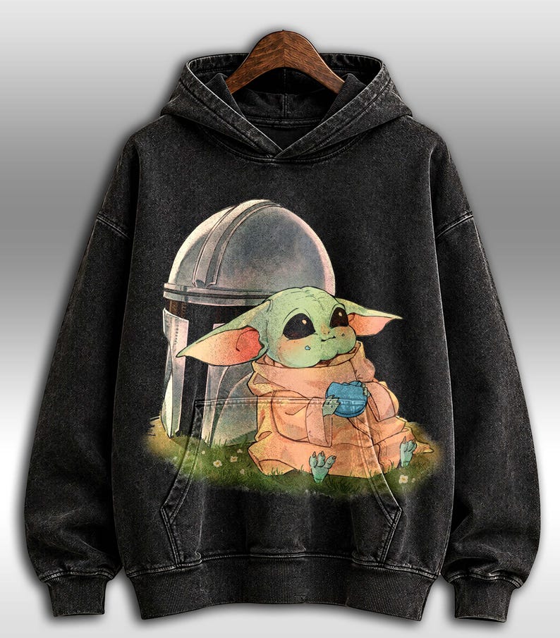 May include: Dark grey hooded sweatshirt with a graphic of Grogu, or Baby Yoda, holding a blue item, with the Mandalorian's helmet behind. The design is printed on a dark, washed-out fabric.