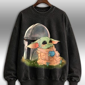 May include: Black crew neck sweatshirt with a graphic of a green alien character holding a blue sphere, with a helmet behind. The alien is wearing a tan robe and sitting on grass. The sweatshirt has a faded, vintage appearance.
