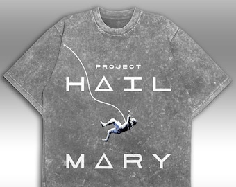 May include: A grey acid-wash short-sleeve t-shirt featuring the white text PROJECT HAIL MARY. A graphic of an astronaut floating in space is positioned between the words, with a thin white curved line extending from the top left corner.