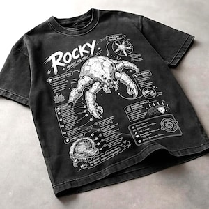 May include: Dark grey t-shirt featuring a white illustration of a crustacean with scientific diagrams. The word "Rocky" is above the illustration, with the text "Project Hail Mary" below. The shirt has a vintage, worn appearance.