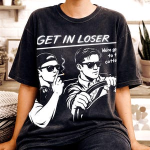 May include: Dark grey t-shirt featuring a graphic of two men in sunglasses, one smoking, the other driving. The text "GET IN LOSER" is above the image, with additional text on the right side.
