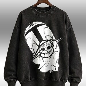 Peut inclure: Sweat-shirt noir &agrave; col rond avec un motif graphique en noir et blanc d'un personnage de dessin anim&eacute;. Le motif comprend un grand "T" et une repr&eacute;sentation stylis&eacute;e d'un personnage avec de grands yeux. Le sweat-shirt a des poignets et un ourlet c&ocirc;tel&eacute;s.
