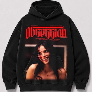 May include: A black hooded sweatshirt featuring a large red graphic print on the chest. The text reads "OBSESSION" in a bold, stylized font above a photographic image of a smiling person with red markings on their face and neck area.