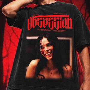 May include: A black short-sleeve t-shirt featuring a graphic print with the word OBSESSION in large red gothic lettering. The design includes a photographic image of a smiling person with blood on their face and text detailing credits.