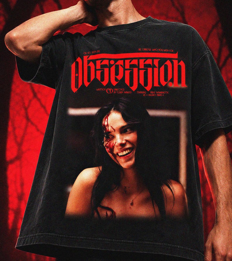 May include: A black short-sleeve t-shirt featuring a graphic print with the word OBSESSION in large red gothic lettering. The design includes a photographic image of a smiling person with blood on their face and text detailing credits.