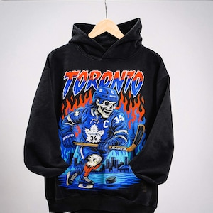 May include: A black hooded sweatshirt featuring a graphic of a skeleton hockey player wearing a blue jersey with the number 34 and a maple leaf logo. The design includes the word TORONTO in blue and orange, flames, and a city skyline background.