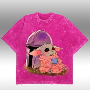 May include: A vibrant pink t-shirt with a cartoon illustration of a character in a pink robe holding a blue item, set against a helmet. The shirt has a textured, faded look.