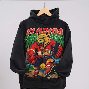 Puede incluir: Sudadera con capucha negra con un gráfico vibrante de un jugador de hockey sobre hielo pantera y la palabra "FLORIDA". La pantera lleva una camiseta roja y equipo de hockey. El diseño incluye palmeras y un atardecer.
