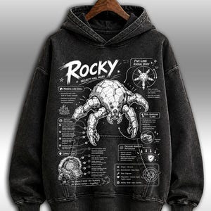 May include: A black hoodie with a detailed graphic design. The design features the word "Rocky", an illustration of a creature, and scientific annotations. The design includes text and diagrams, creating a unique aesthetic.