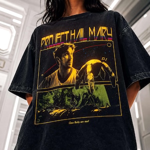 Puede incluir: Una camiseta negra con efecto lavado al ácido que presenta un estampado gráfico con el texto "PROJECT HAIL MARY" en la parte superior. El diseño incluye el retrato de un hombre, una criatura arácnida y el texto "Grace Rocky save stars!".