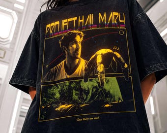 May include: A black acid-wash t-shirt featuring a graphic print with the text "PROJECT HAIL MARY" at the top. The design includes a portrait of a man and a multi-legged creature, with the text "Grace Rocky save stars!" at the bottom.