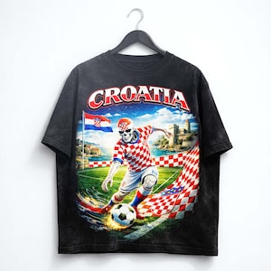 May include: Black t-shirt featuring a soccer-playing skeleton in a Croatian team uniform. The design includes the word "CROATIA" in red and white, a Croatian flag, and a castle in the background. The shirt is hanging on a black hanger.