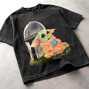May include: Black t-shirt with a graphic of a green alien character from The Mandalorian, with a helmet behind it. The alien is holding a blue object and wearing a tan robe. The shirt has a vintage wash.