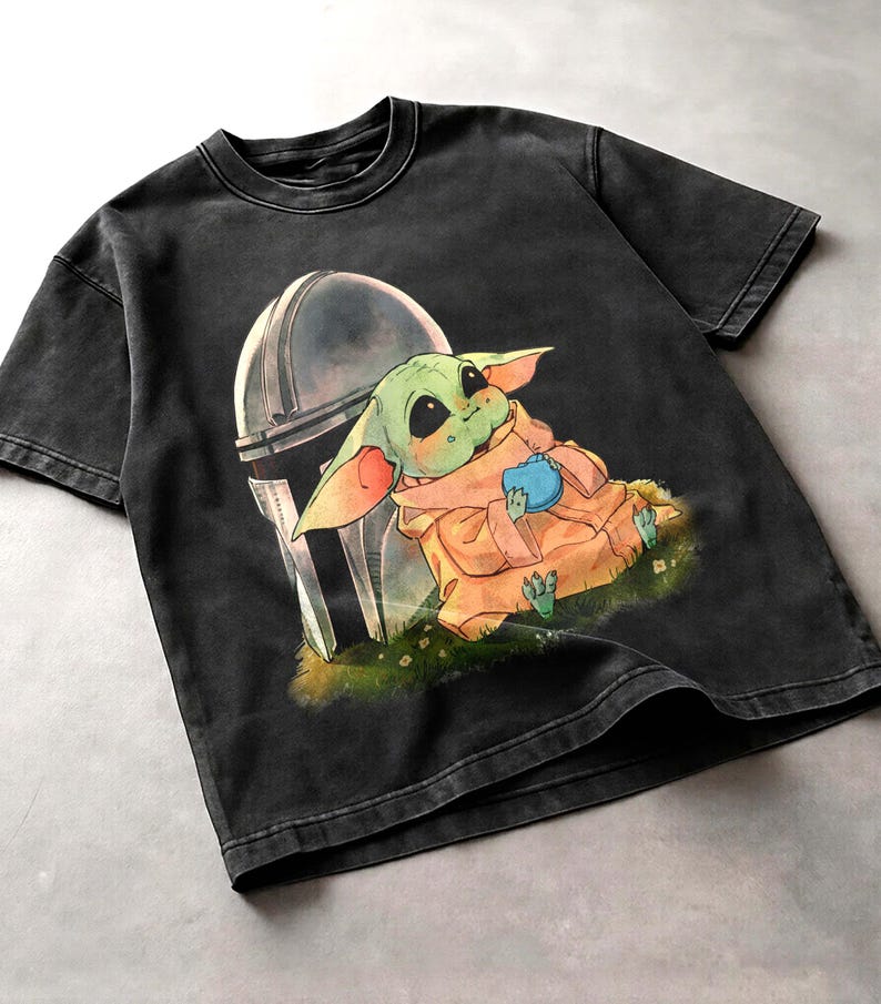 May include: Black t-shirt with a graphic of a green alien character from The Mandalorian, with a helmet behind it. The alien is holding a blue object and wearing a tan robe. The shirt has a vintage wash.
