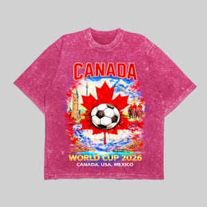 May include: A magenta t-shirt with a graphic design featuring a soccer ball on a red maple leaf, the word "CANADA" in red, and the text "WORLD CUP 2026 CANADA, USA, MEXICO". The design includes a landscape with buildings and figures.
