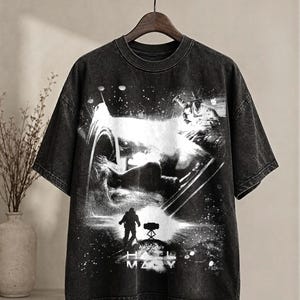 May include: Black graphic t-shirt with a black and white space-themed illustration, including a figure and the text "H. MZ". The tee is displayed on a black clothes rail.