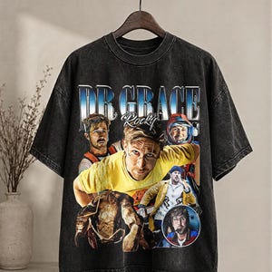 May include: Dark grey vintage-style t-shirt with a graphic design featuring the text "DR GRACE Rocky" in a blue and white gradient font. The design includes multiple portraits of a person. The shirt has a crew neck and short sleeves.