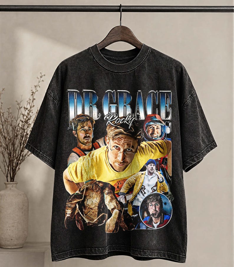 May include: Dark grey vintage-style t-shirt with a graphic design featuring the text "DR GRACE Rocky" in a blue and white gradient font. The design includes multiple portraits of a person. The shirt has a crew neck and short sleeves.