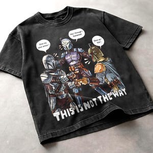 K&ouml;nnte beinhalten: Schwarzes Grafik-T-Shirt mit einer Illustration zum Thema Star Wars. Das Design zeigt drei Charaktere in mandalorianischer R&uuml;stung mit Sprechblasen. Der Text auf dem Shirt lautet "THIS IS NOT THE WAY".