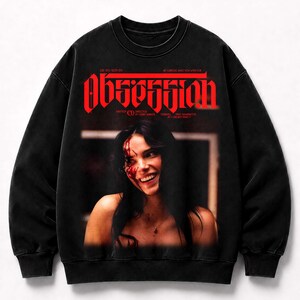 May include: A black crewneck sweatshirt featuring a graphic print of a smiling person with blood on their face. The word "OBSESSION" is printed in large red letters above the image, with additional small text details included in the design.