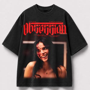 May include: A black short-sleeve t-shirt featuring a graphic print of a smiling person with blood on their face. The word "OBSESSION" is printed in large red letters at the top, with additional small text including "WRITTEN" and "DIRECTED".