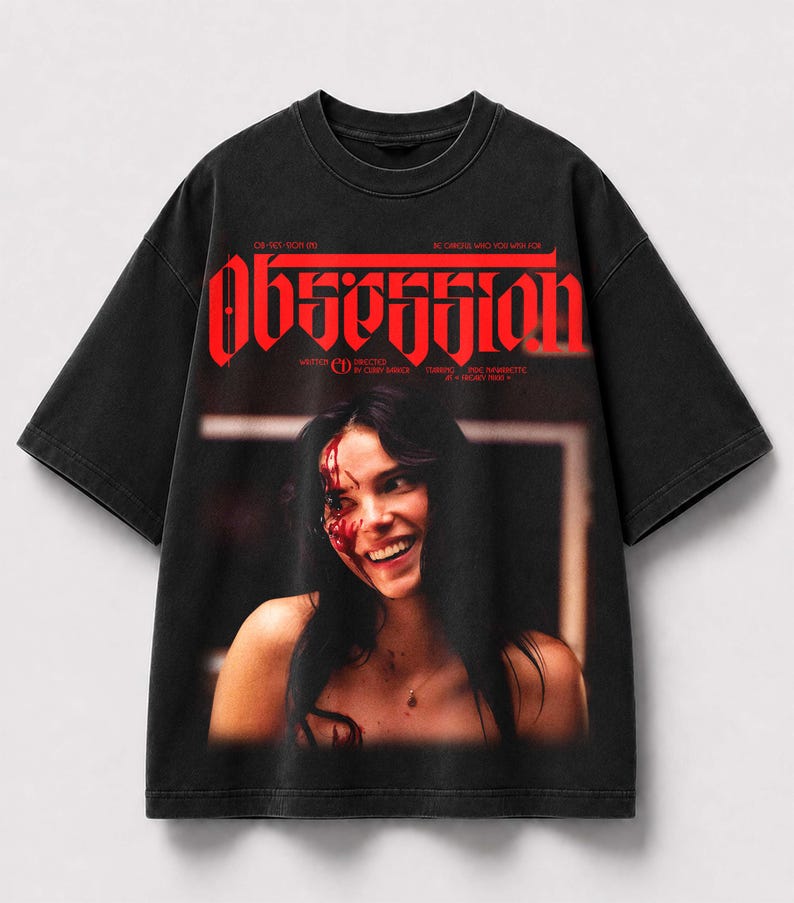 May include: A black short-sleeve t-shirt featuring a graphic print of a smiling person with blood on their face. The word "OBSESSION" is printed in large red letters at the top, with additional small text including "WRITTEN" and "DIRECTED".