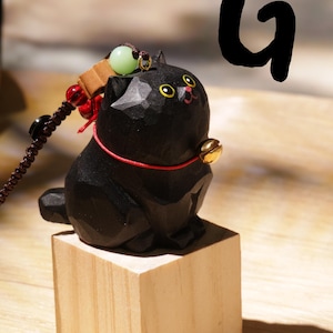 May include: A carved black cat figurine with yellow eyes and a red collar, sitting on a small wooden block. The cat has a gold bell and a decorative string with beads. The letter "G" is in the background.