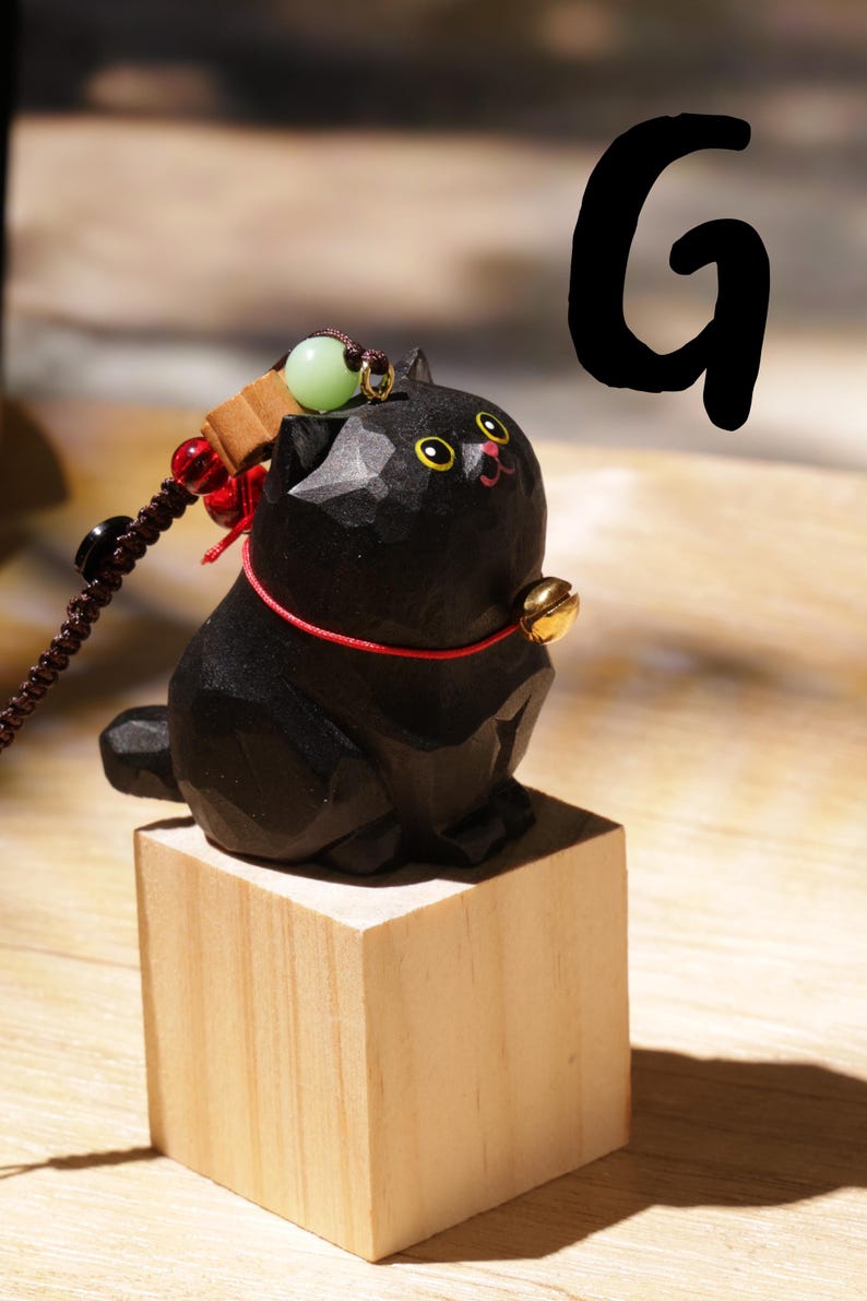 May include: A carved black cat figurine with yellow eyes and a red collar, sitting on a small wooden block. The cat has a gold bell and a decorative string with beads. The letter "G" is in the background.