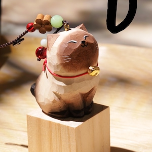 May include: A carved wooden cat figurine with a gold bell collar and red string. The cat is light brown with darker brown accents and sits on a small wooden block. A decorative string with beads and a flower is attached.