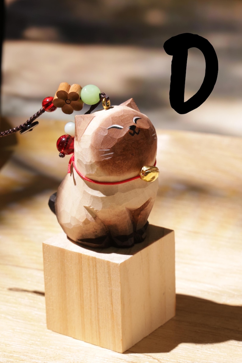 May include: A carved wooden cat figurine with a gold bell collar and red string. The cat is light brown with darker brown accents and sits on a small wooden block. A decorative string with beads and a flower is attached.