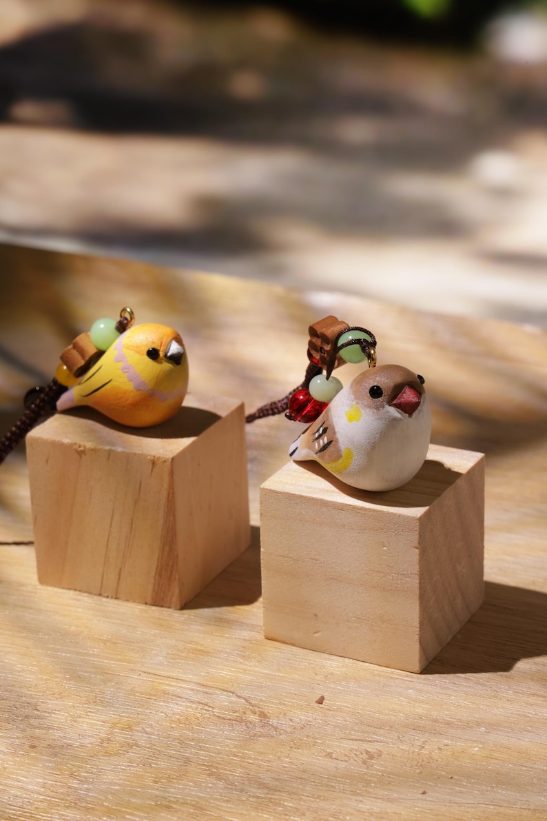 May include: Two small bird-shaped charms, one yellow and one brown and white, each featuring a dark beak and eyes. The charms are attached to dark brown cords with small decorative beads in red, green, and brown, resting on two light wood cubes.