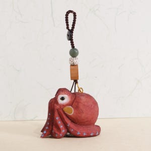 May include: A hand-carved, red octopus ornament with blue star accents on its tentacles. The octopus is suspended by a brown cord with a wooden bead, a speckled bead, and a square wooden piece. A small gold-colored hook connects the octopus to the cord.