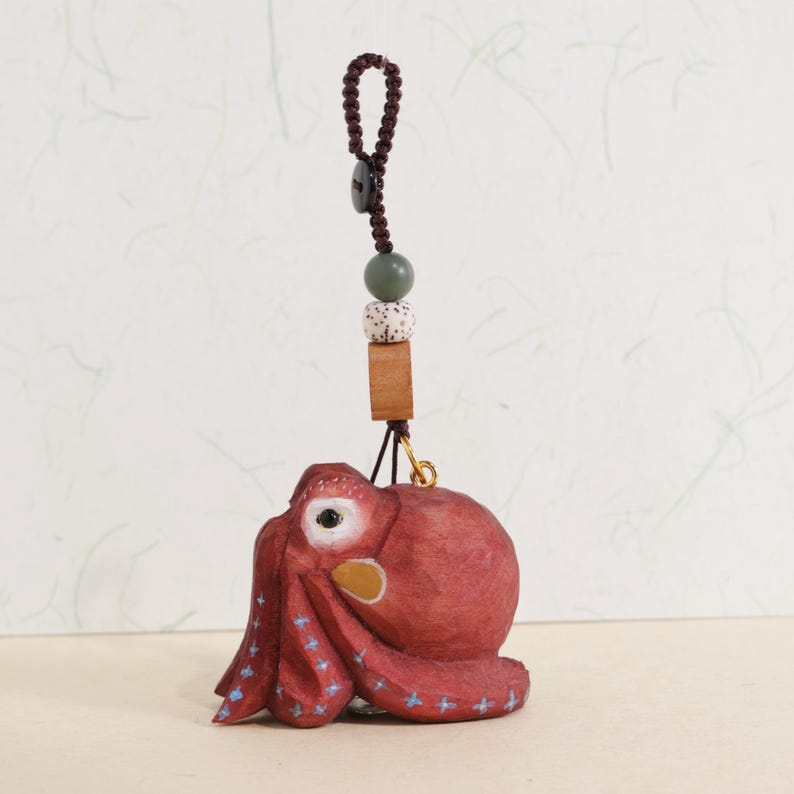 May include: A hand-carved, red octopus ornament with blue star accents on its tentacles. The octopus is suspended by a brown cord with a wooden bead, a speckled bead, and a square wooden piece. A small gold-colored hook connects the octopus to the cord.