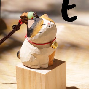 May include: A wooden cat figurine with a calico pattern, sitting on a small wooden block. The cat has a red collar with a gold bell. A decorative cord with beads is attached to the cat's head.