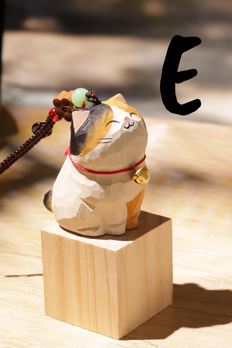 May include: A wooden cat figurine with a calico pattern, sitting on a small wooden block. The cat has a red collar with a gold bell. A decorative cord with beads is attached to the cat's head.