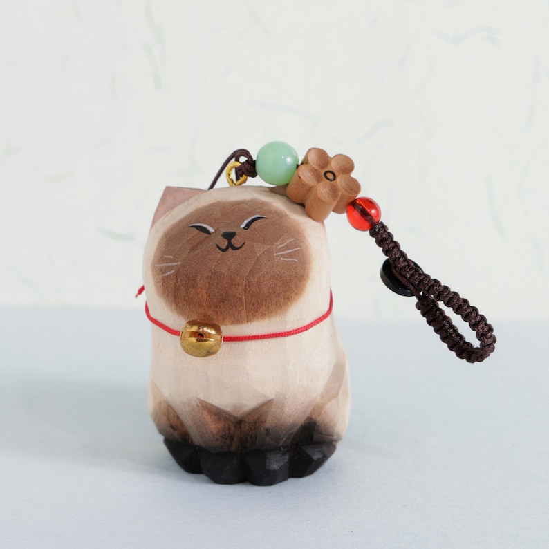 May include: A wooden cat figurine with a smiling face, wearing a red collar with a gold bell. The cat has brown and beige coloring, with a brown cord and decorative beads attached. The figurine is a charming decorative item.
