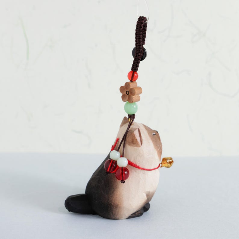 May include: A handcrafted wooden cat figurine with a bell, adorned with a decorative cord and beads. The cat is primarily brown and cream-colored, with a small gold bell. The cord features a flower-shaped bead and other colorful accents.