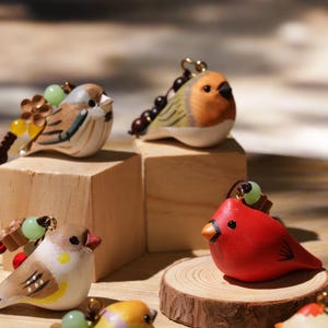 Può includere: Una collezione di figurine di uccelli dipinte a mano in vari colori, tra cui rosso, giallo e marrone. Ogni uccello ha un design dettagliato ed è attaccato a una corda di perline decorativa. Alcuni uccelli sono esposti su blocchi di legno e una fetta di legno.