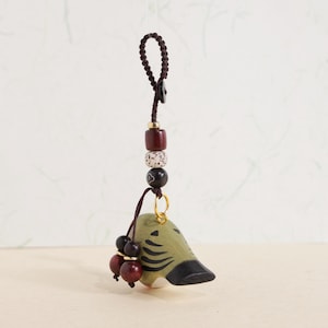 May include: A handcrafted, decorative charm featuring a bird-like figure in olive green with black accents. The charm is suspended from a dark brown cord with a series of beads in various colors and textures, including red, white, and black. A small gold ring connects the charm to the cord.