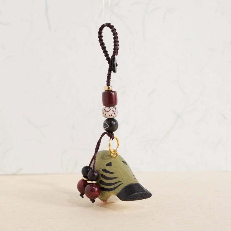 May include: A handcrafted, decorative charm featuring a bird-like figure in olive green with black accents. The charm is suspended from a dark brown cord with a series of beads in various colors and textures, including red, white, and black. A small gold ring connects the charm to the cord.