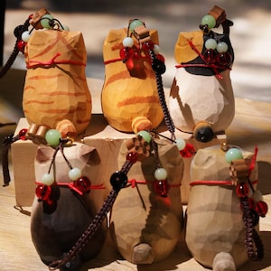 May include: A collection of six carved wooden cat figurines. Each cat has a unique color and pattern, with details like red string and beaded accents. The figurines are displayed on a wooden surface, showcasing their handcrafted nature.