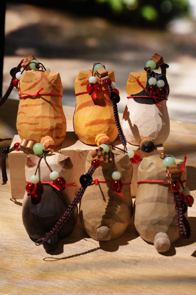 May include: A collection of six carved wooden cat figurines. Each cat has a unique color and pattern, with details like red string and beaded accents. The figurines are displayed on a wooden surface, showcasing their handcrafted nature.