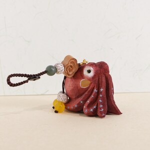 May include: A hand-carved, red octopus keychain with a brown braided loop. The octopus has white star accents and a yellow bead. The keychain also features wooden and speckled beads.
